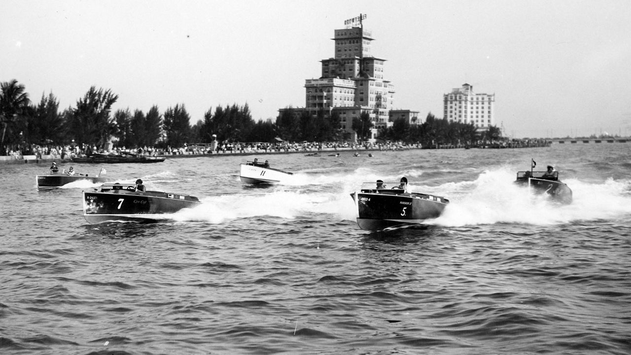 Multiple chris craft boats on the water with buildings in the background