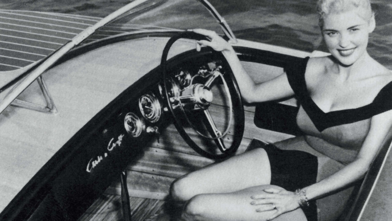 A lady sitting in the drivers seat of a chris craft boat