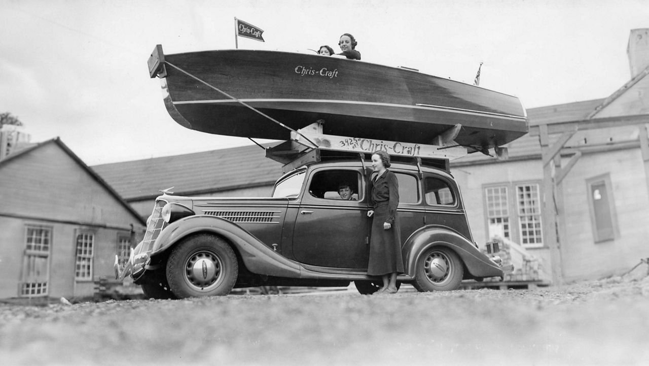 An old car with a chris craft boat strapped to the top