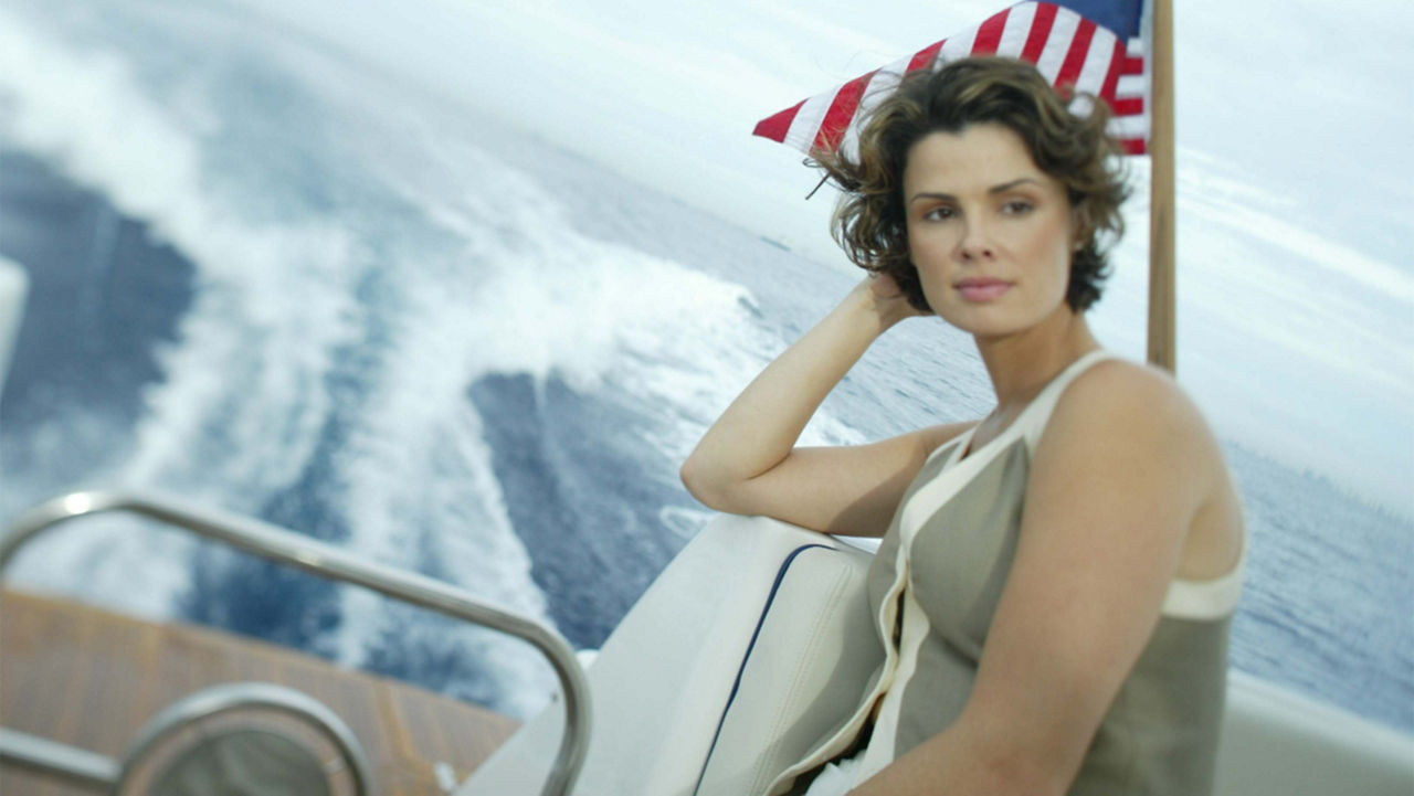 A woman looking off to the right sitting in a chris craft boat with the ocean and an american flag behind her